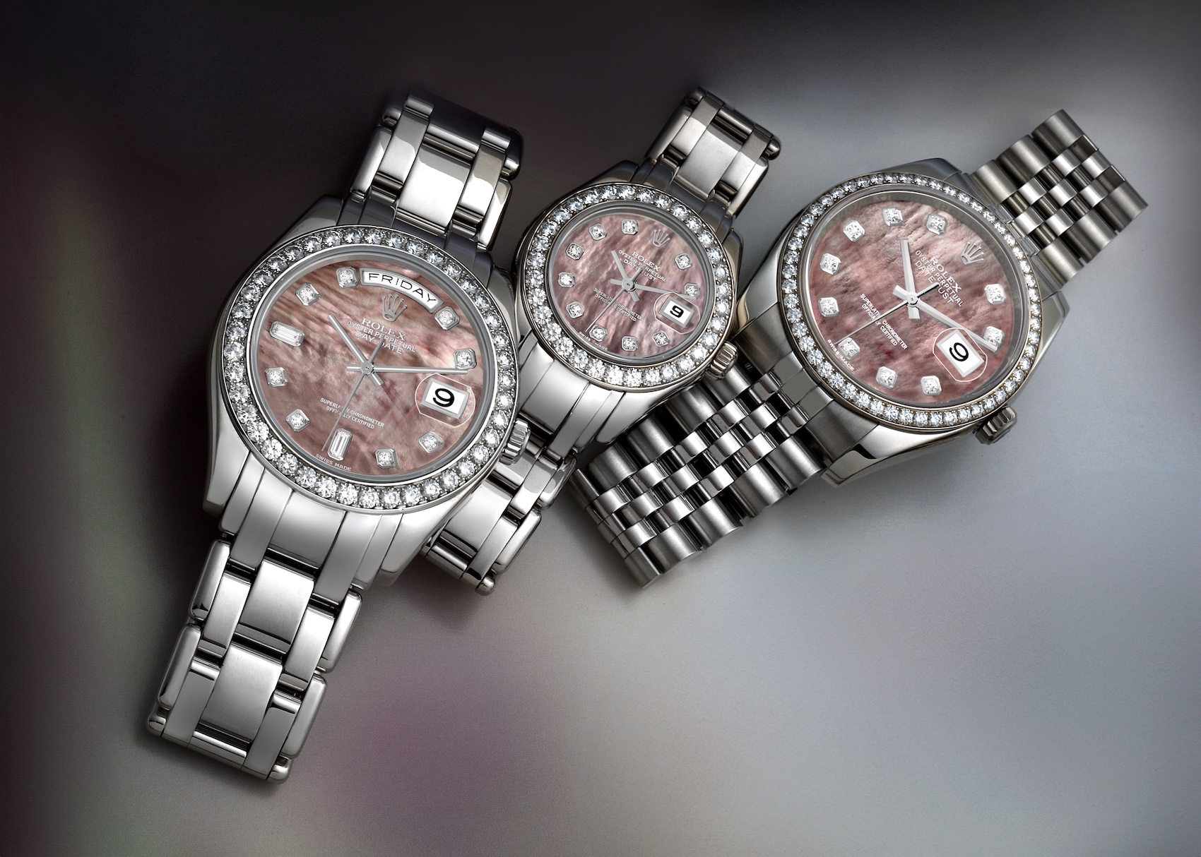 Three luxury Rolex watches in white gold or platinum, each featuring a diamond-set bezel and a unique pink or lavender mother-of-pearl dial, displayed on a grey surface.