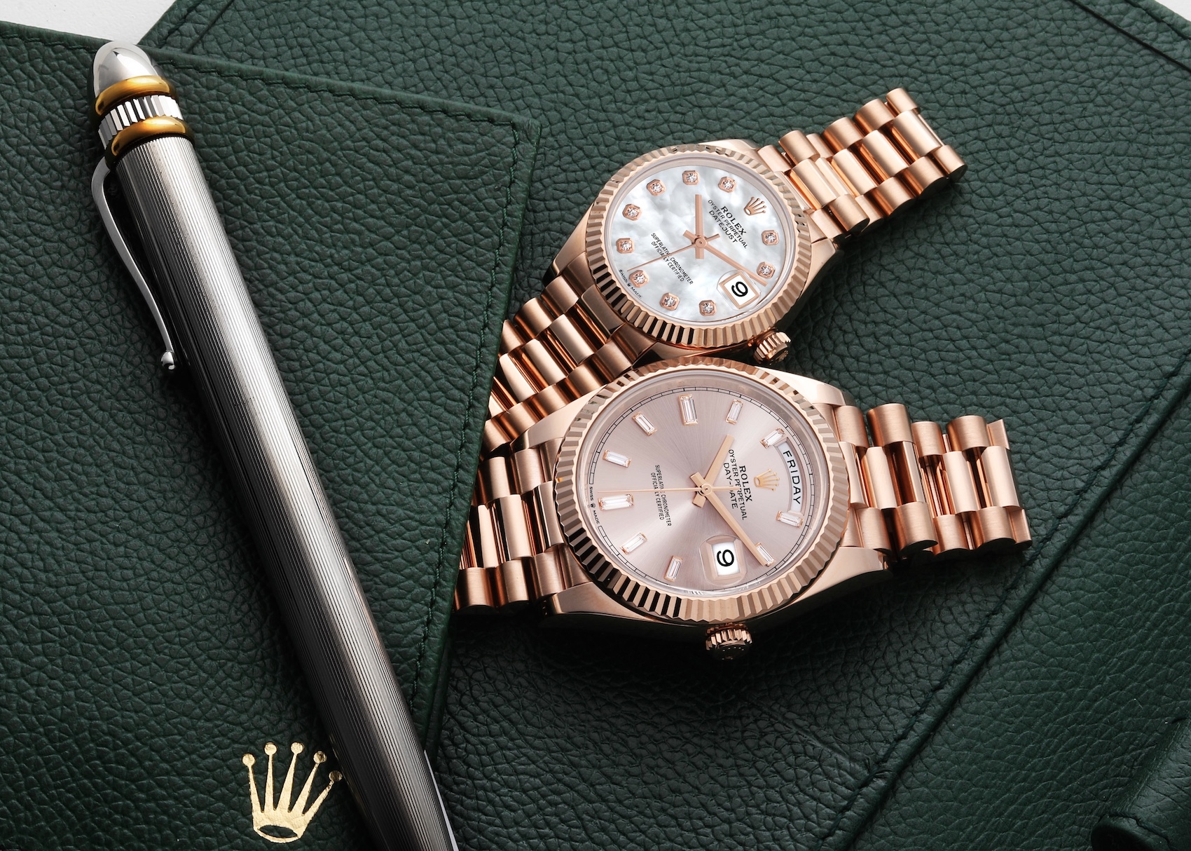 Two rose gold Rolex watches, one Day-Date with a champagne dial and a Datejust with a diamond-set mother-of-pearl dial, lying on a dark green leather surface next to a pen.