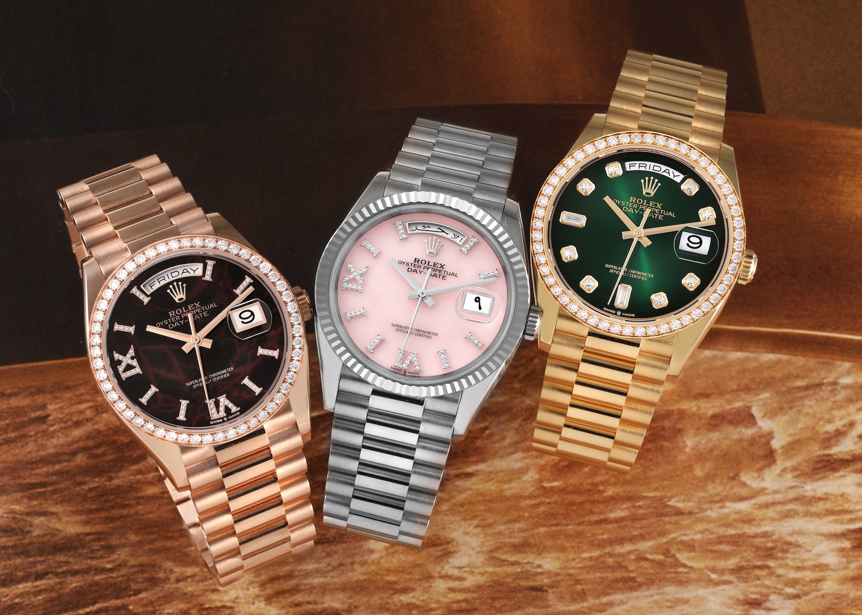 Three luxury Rolex Day-Date watches, including rose gold with a dark dial, white gold with a pink dial, and yellow gold with a green diamond-set dial, all featuring diamond bezels.