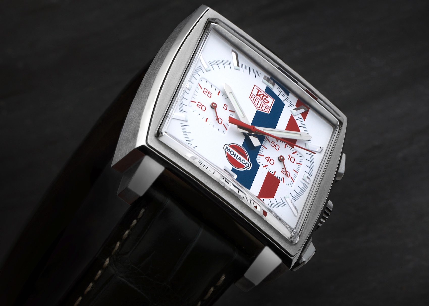 An angled close-up of the square-cased TAG Heuer Monaco Gulf CW2118 watch, featuring a white dial with iconic red and blue Gulf stripes and a black leather strap.