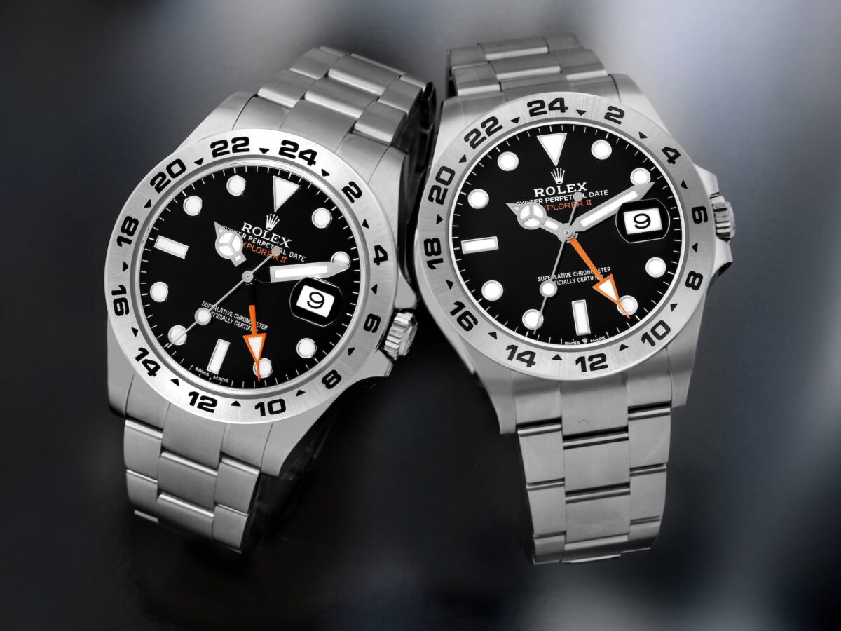 Two stainless steel Rolex Explorer II watches are shown side-by-side on a dark surface: on the left, the older reference 216570 with a 42mm case, and on the right, the newer reference 226570 with subtle refinements to the case and bracelet. Both feature black dials, white luminous markers, orange 24-hour hands, and fixed 24-hour bezels.