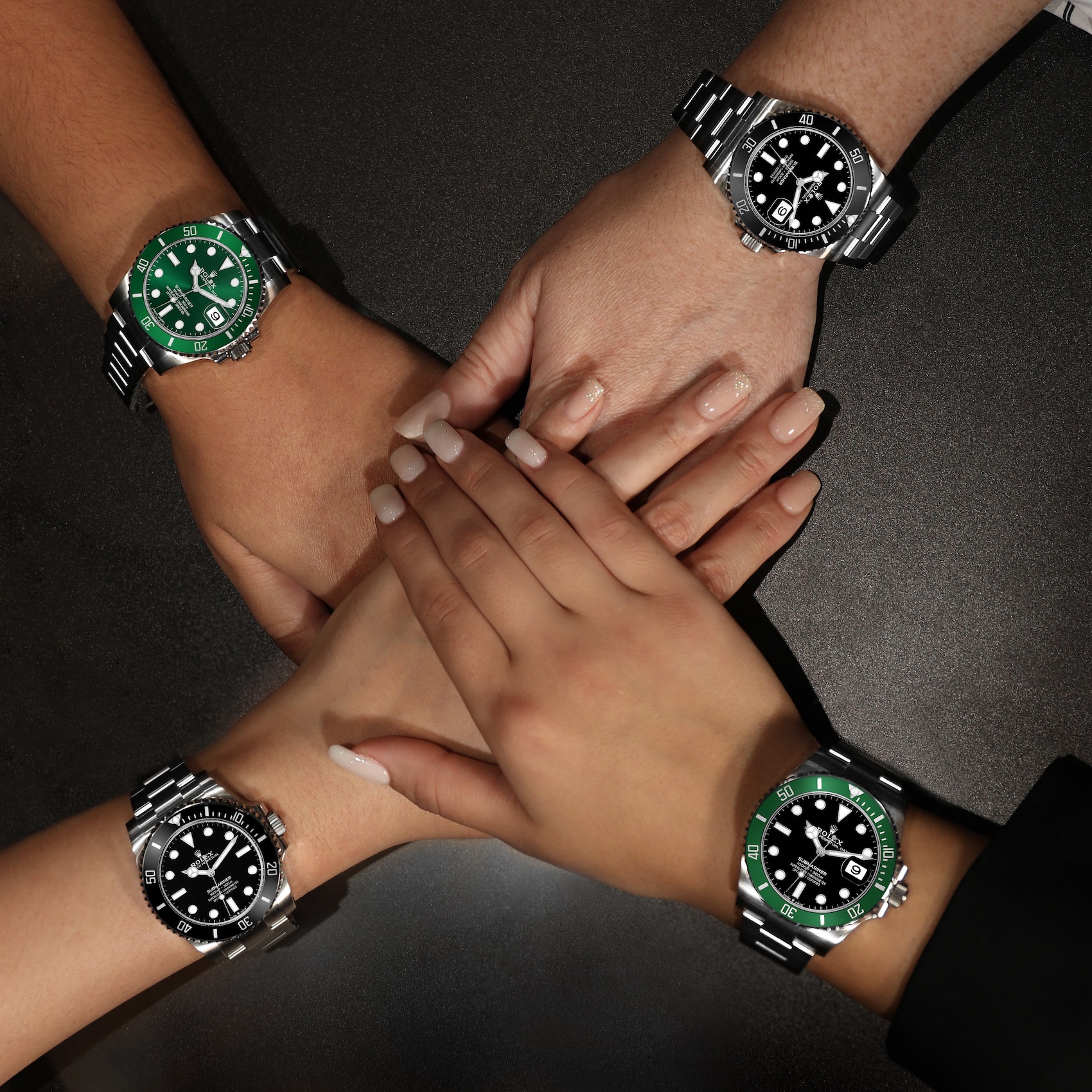 Four wrists are stacked, each wearing a Rolex Submariner. The top and bottom wrists wear a green bezel Submariner and the middle wrists wear a black bezel Submariner.