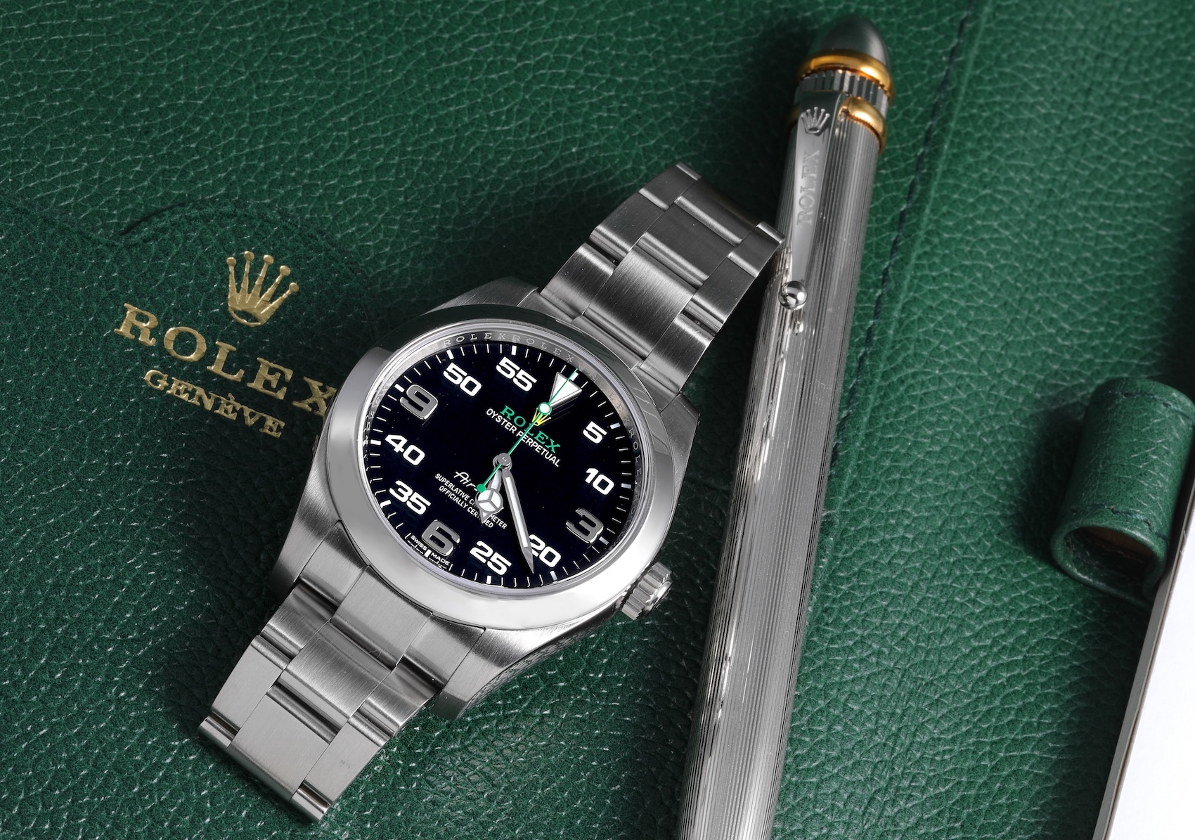 A stainless steel Rolex Oyster Perpetual Air-King reference 116900 with a black dial featuring a distinctive aviation-inspired minute scale and the Rolex and Air-King logos in green and yellow is positioned next to a silver Rolex pen on a green Rolex document holder.