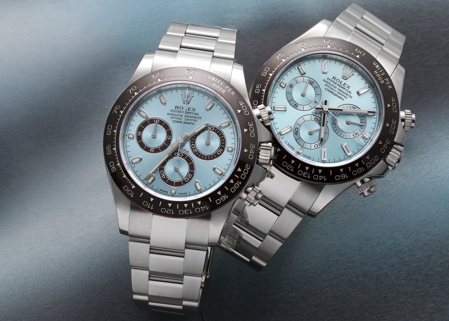 Two platinum Rolex Daytona watches with ice-blue dials and chestnut brown Cerachrom bezels are positioned closely together on a textured grey surface.