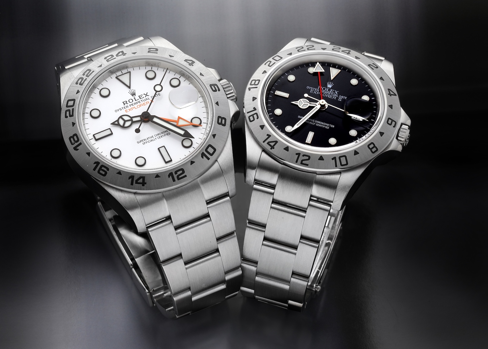 Two stainless steel Rolex Explorer II watches are displayed on a dark, reflective surface: on the left, a reference 216570 with a white "Maxi" dial and orange 24-hour hand, and on the right, a reference 16570 with a black dial and red 24-hour hand.