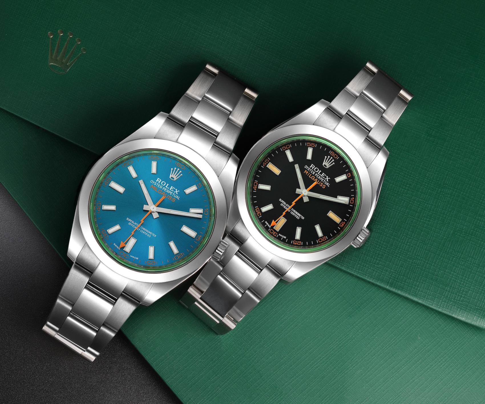 Two stainless steel Rolex Milgauss 116400 watches are positioned on a green Rolex watch box: one with a vibrant Z-blue dial and the other with a black dial, both featuring the signature orange lightning bolt seconds hand and a green-tinted crystal edge.
