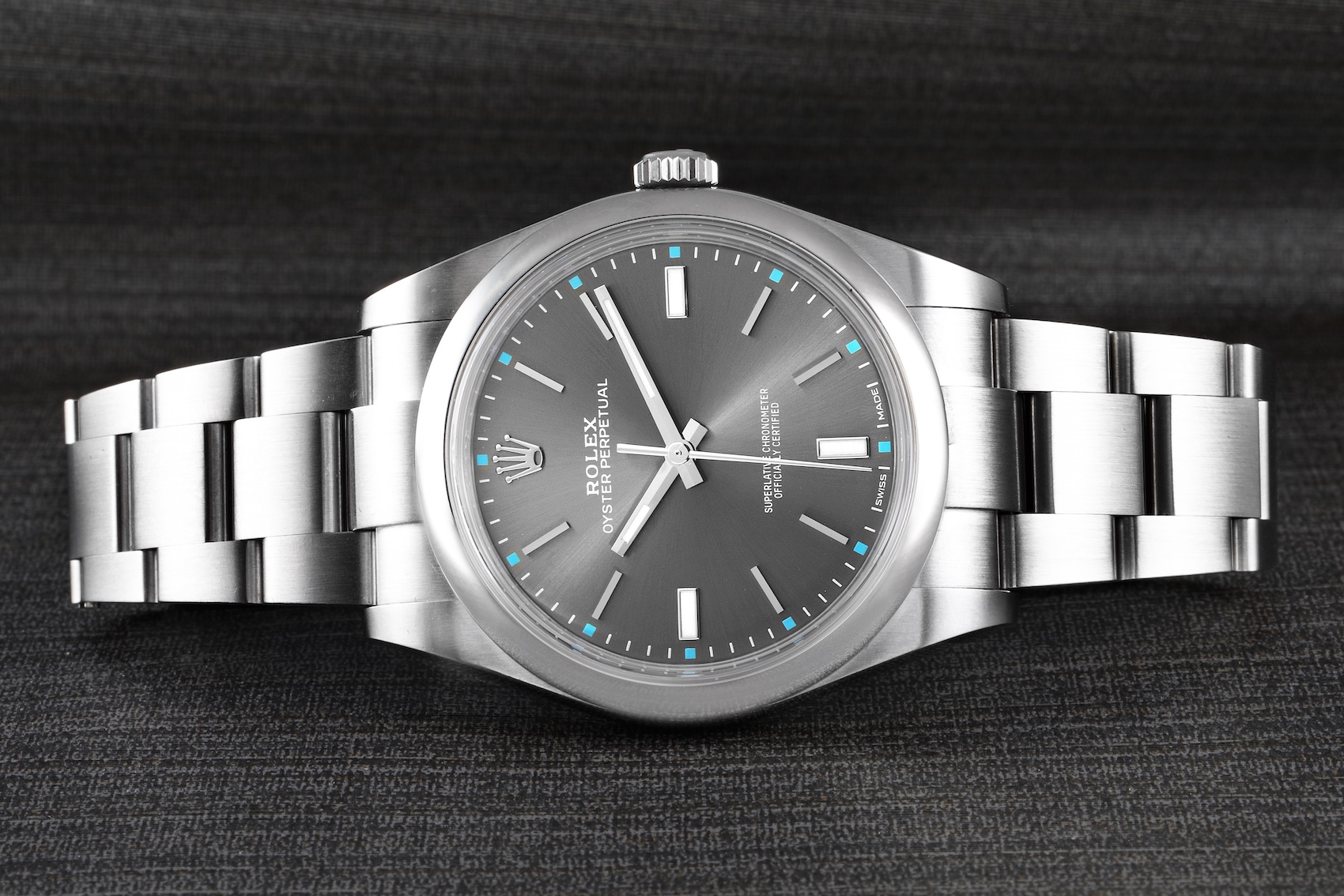 A stainless steel Rolex Oyster Perpetual 39 with a rhodium dial and light blue accents is displayed on a dark, textured surface, showcasing its clean lines and minimalist design.