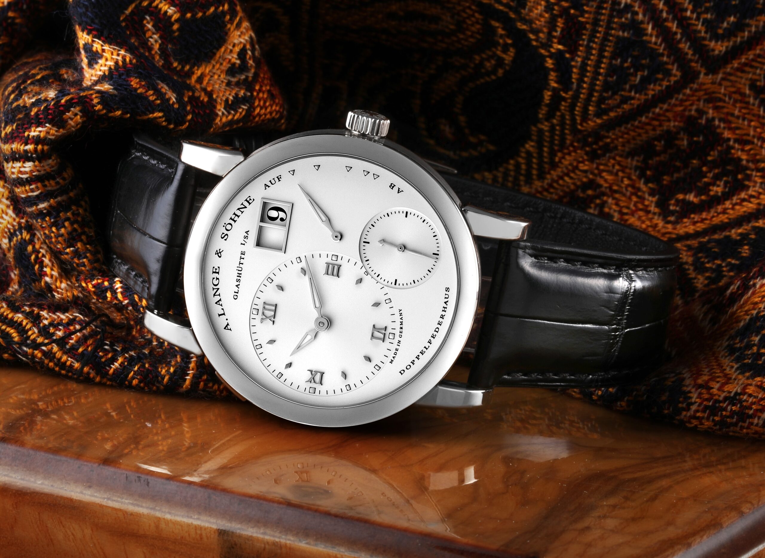 The iconic A. Lange & Söhne Lange 1 in white gold, with its classic silver dial and asymmetrical layout, resting on a textured, patterned fabric.