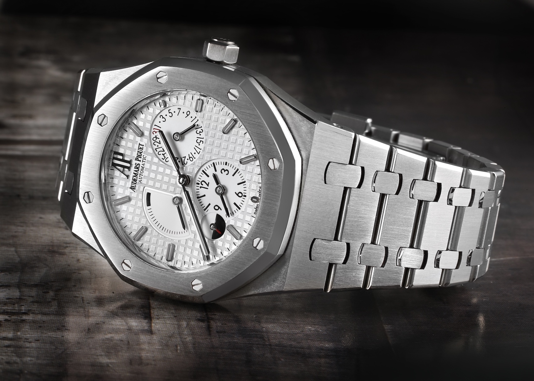 An Audemars Piguet Royal Oak Dual Time watch on a dark, textured surface. It features the signature octagonal bezel with exposed screws, a silver-toned dial with a 'Grande Tapisserie' pattern, and an integrated steel bracelet.