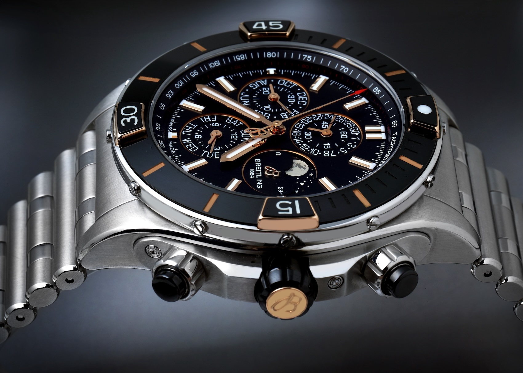 Close-up angled view of a Breitling Super Chronomat Four Year Calendar watch. It features a stainless steel case, a black ceramic bezel with 18k red gold rider tabs, a complex black dial with multiple subdials and a moonphase, and a steel Rouleaux bracelet.