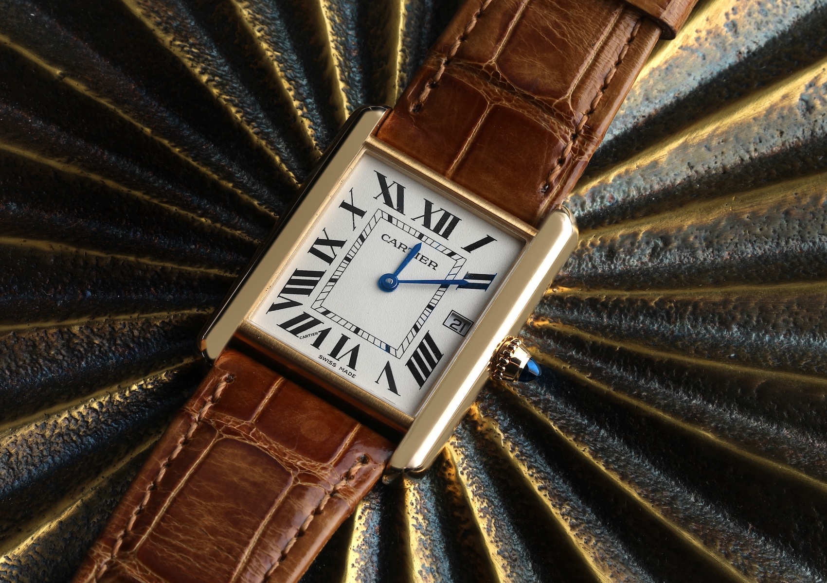 The iconic Cartier Tank Louis watch shown with its rectangular yellow gold case, classic white dial, and a brown alligator leather strap, resting on a textured gold surface.