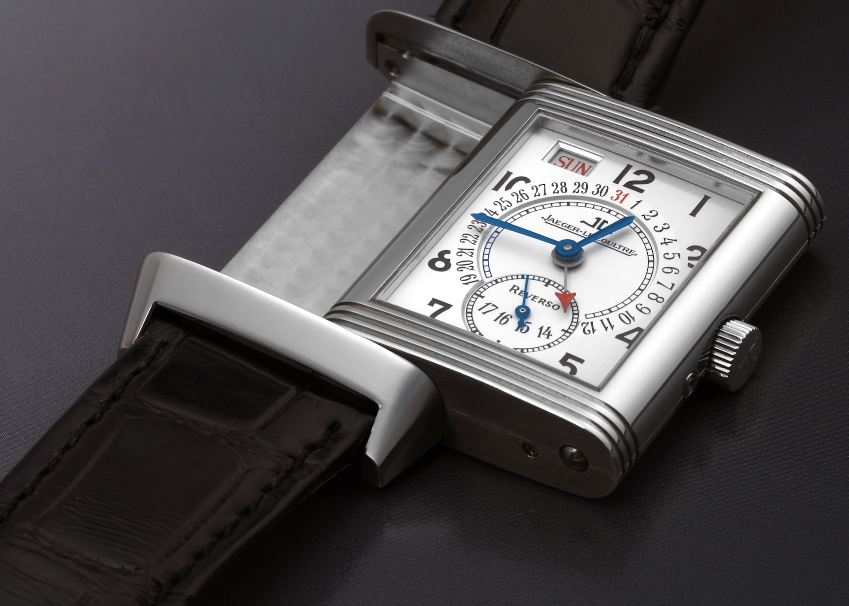 A Jaeger-LeCoultre Grande Reverso watch with its case partially swiveled out of its carrier. The watch has a rectangular steel case, a silvered guilloché dial with multiple complications, and a black alligator strap.