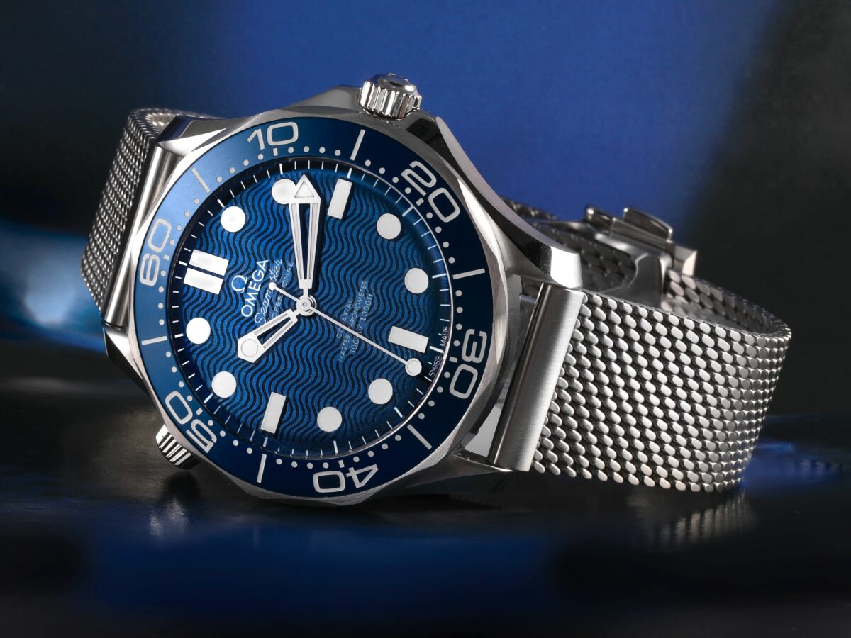 A modern OMEGA Seamaster Diver 300M with a blue ceramic wave dial, shown on a steel Milanese mesh bracelet.