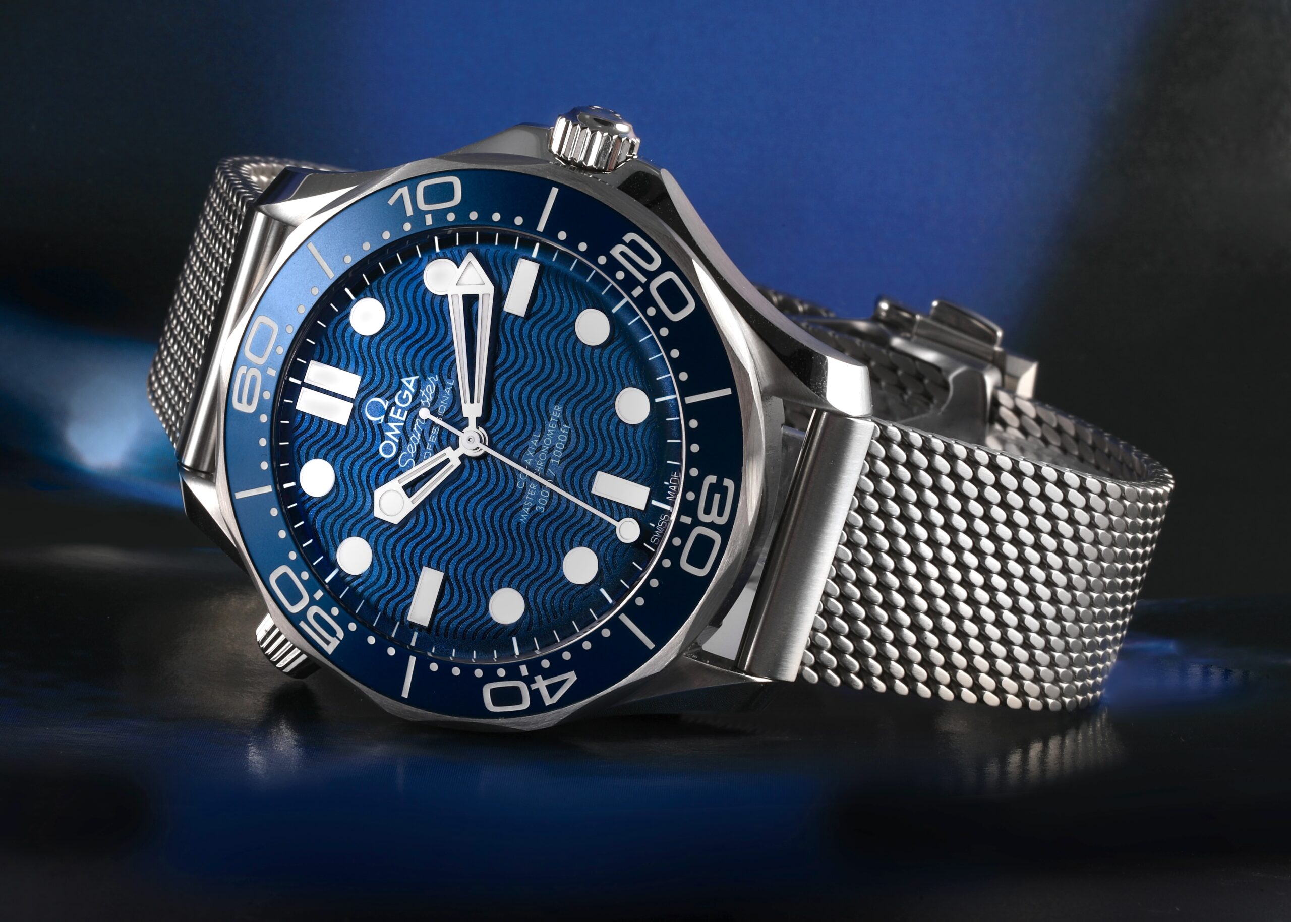 A modern OMEGA Seamaster Diver 300M with a blue ceramic wave dial, shown on a steel Milanese mesh bracelet.