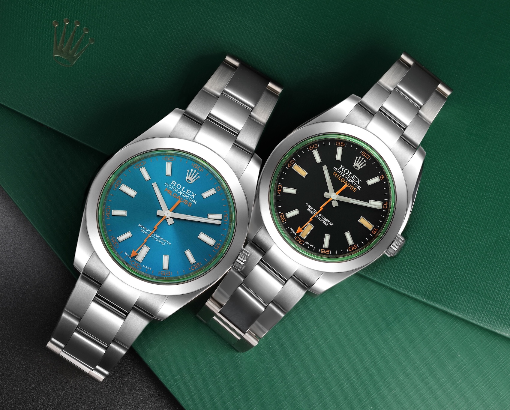 Two Rolex Milgauss 116400GV watches side-by-side on a green background. The left watch has a Z-Blue dial, and the right has a black dial. Both feature the signature orange lightning-bolt seconds hand and green-tinted sapphire crystal.
