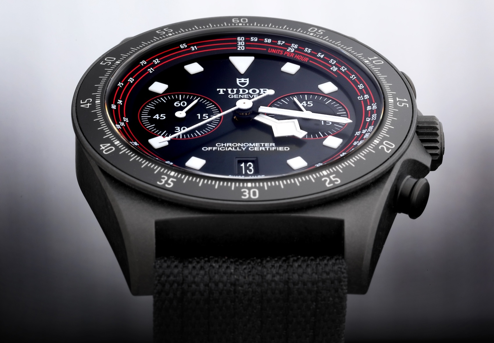 A Tudor Pelagos FXD Chrono Cycling Edition watch shown from an angle. The watch has a matte black carbon composite case, a black dial with two subdials and red accents, and a black fabric strap.
