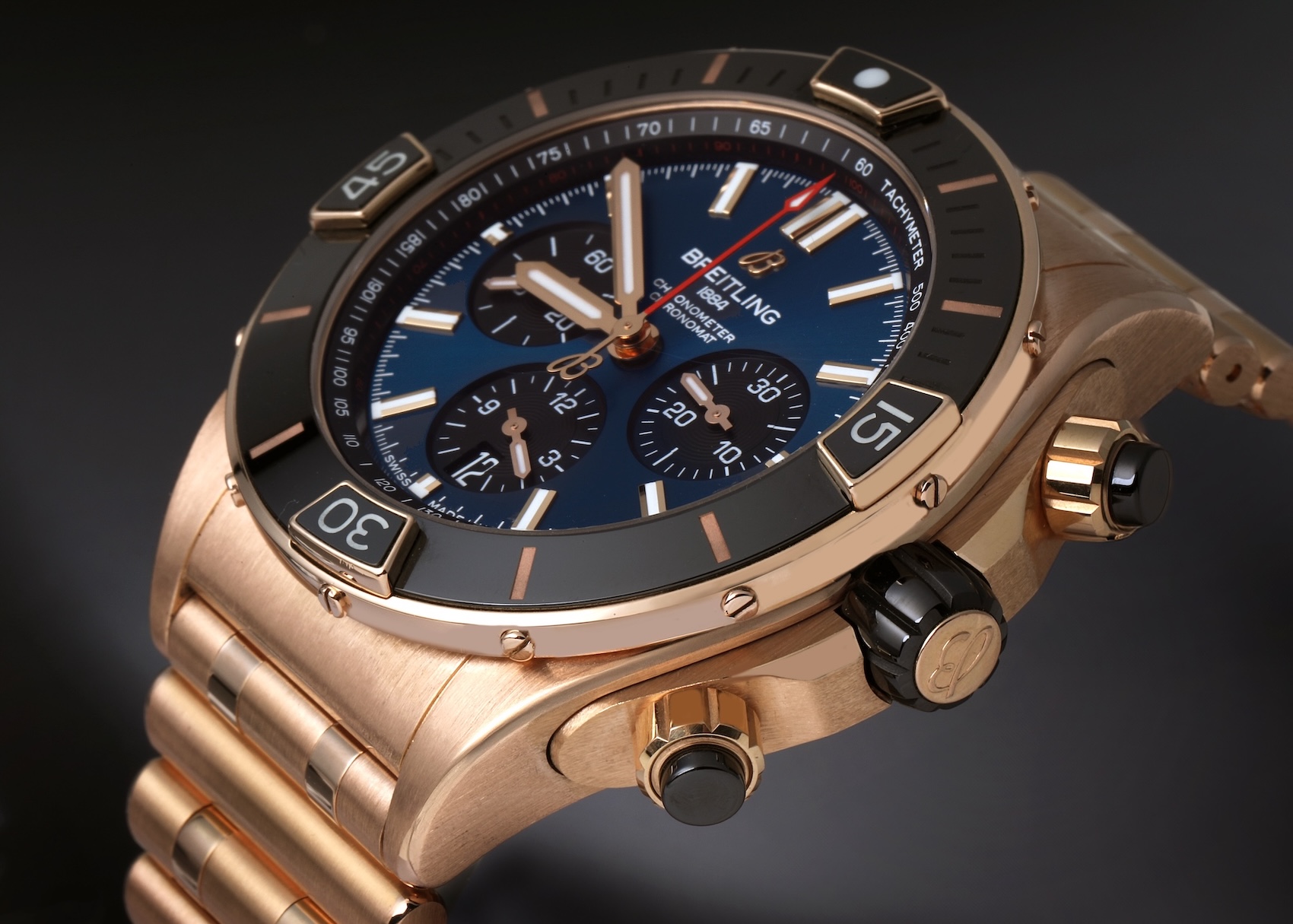 An angled view of a luxurious Breitling Chronomat B01 watch in 18k rose gold, featuring a deep blue dial, black ceramic bezel with gold rider tabs, and black pushers.