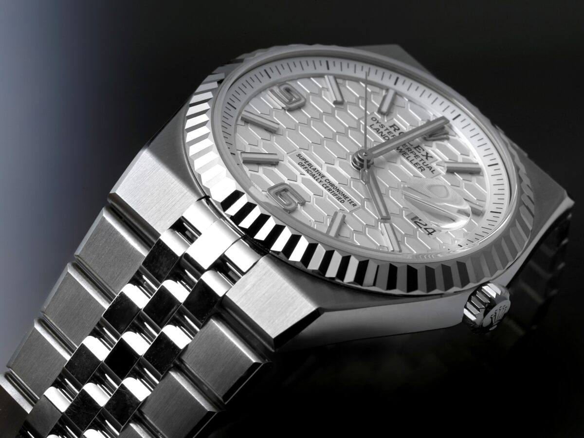 A close-up, angled view of the Rolex Land-Dweller watch in dramatic lighting. The image highlights the fluted bezel, the white honeycomb dial, and the Jubilee-style bracelet against a dark, reflective surface.