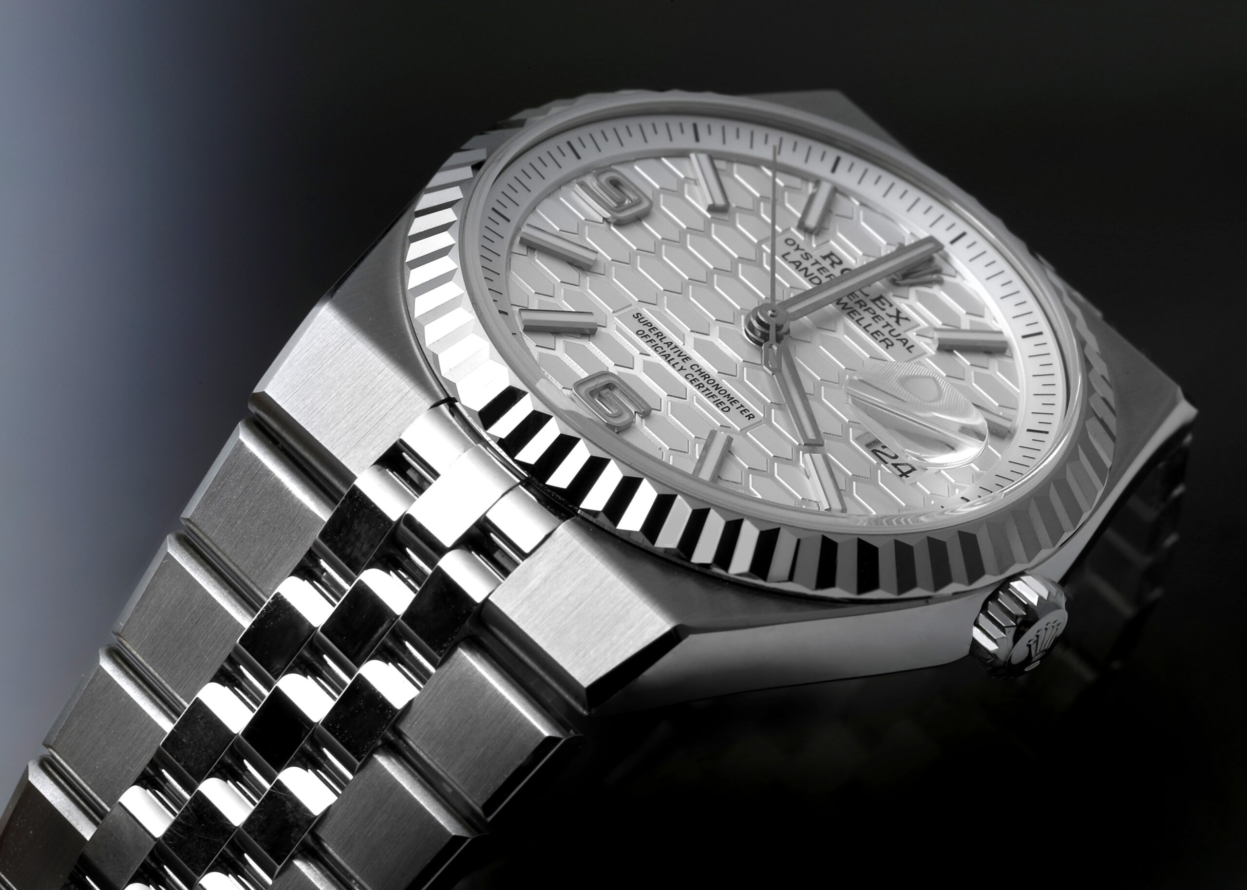 A close-up, angled view of the Rolex Land-Dweller watch in dramatic lighting. The image highlights the fluted bezel, the white honeycomb dial, and the Jubilee-style bracelet against a dark, reflective surface.