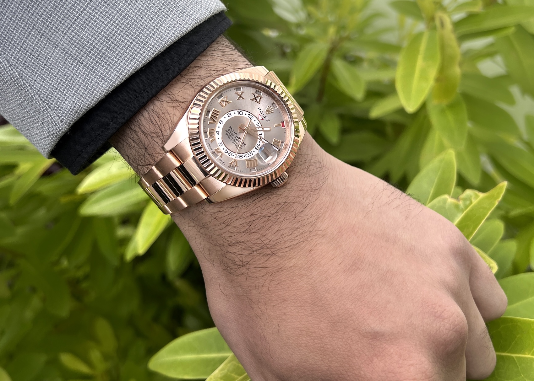 A man wearing an Everose gold Rolex Sky-Dweller with a sundust dial and Roman numerals on his wrist, shown outdoors against a background of green leaves.