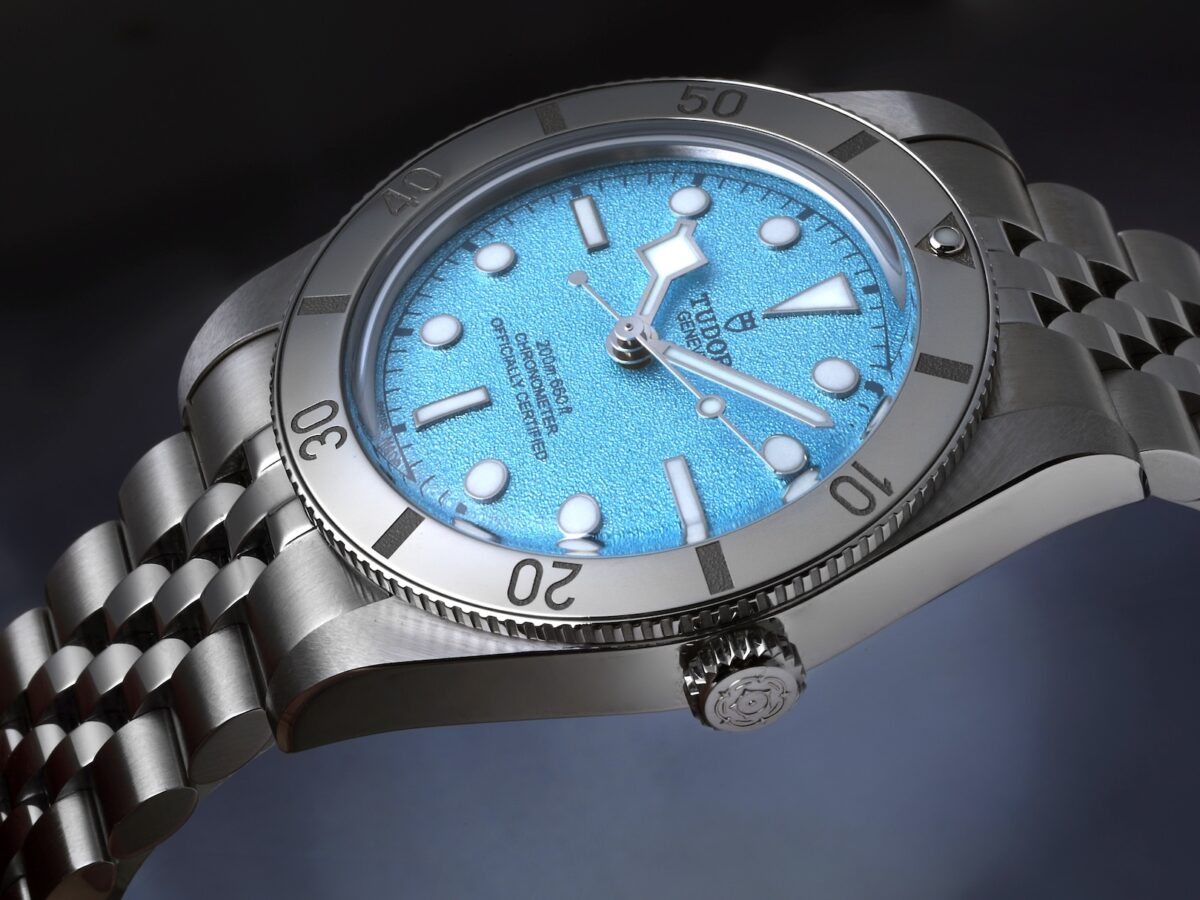 A stylish three-quarter view of the Tudor Black Bay 54, emphasizing the brushed steel case, the five-link bracelet, and the prominent screw-down crown engraved with the Tudor rose logo.