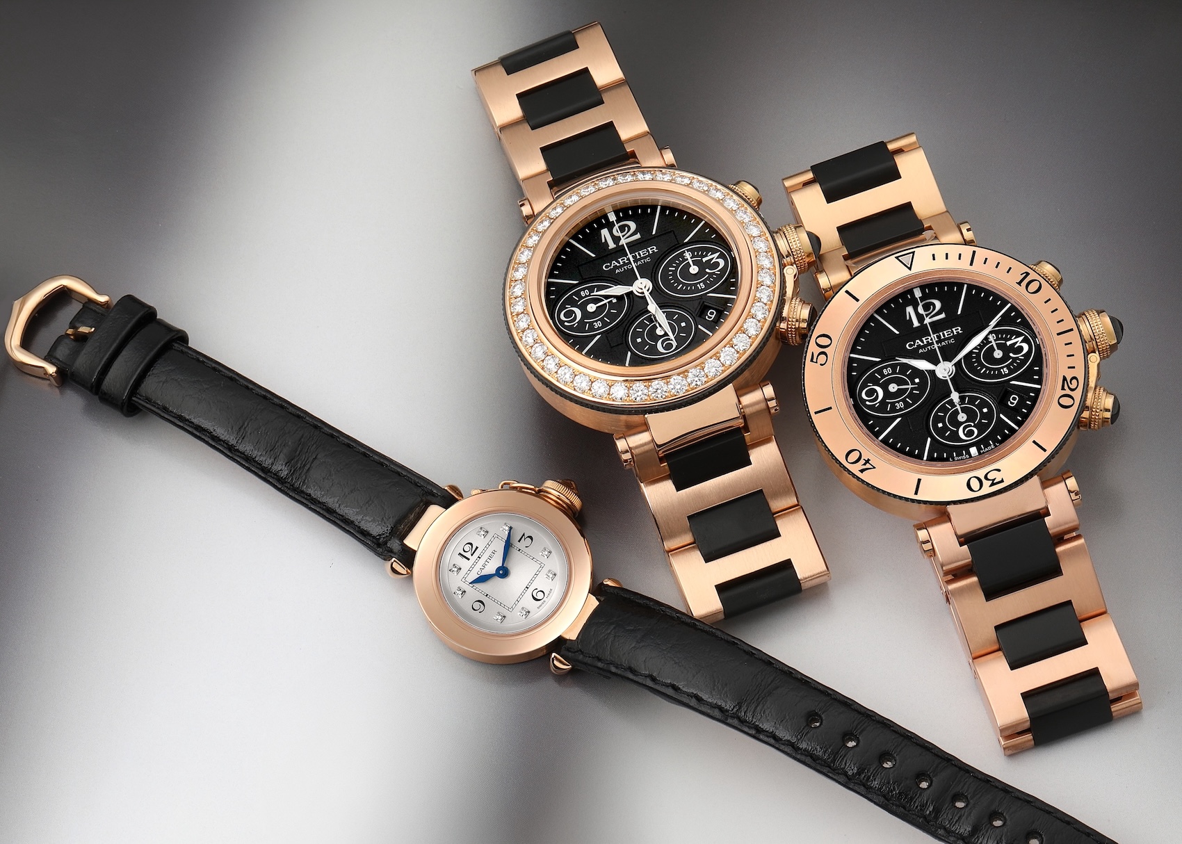 A group of three Cartier Pasha watches in 18k rose gold. Two are large Pasha Seatimer chronographs with black dials, one with a diamond bezel, and the third is a smaller Pasha 27 ladies' watch with a white dial.