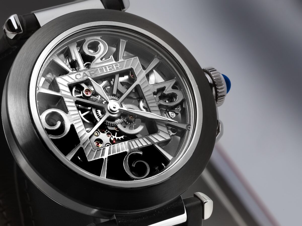 A detailed macro shot of a Cartier Pasha de Cartier Skeleton watch. The open-worked movement forms the Arabic numerals, and the iconic chained crown with its blue cabochon is visible.