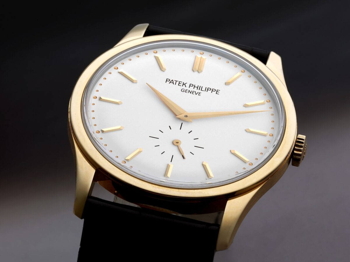 A close-up of a Patek Philippe Calatrava ref 5196J in yellow gold. The watch has a silver dial with gold baton hour markers, dauphine hands, and a small seconds sub-dial at 6 o'clock on a black strap.
