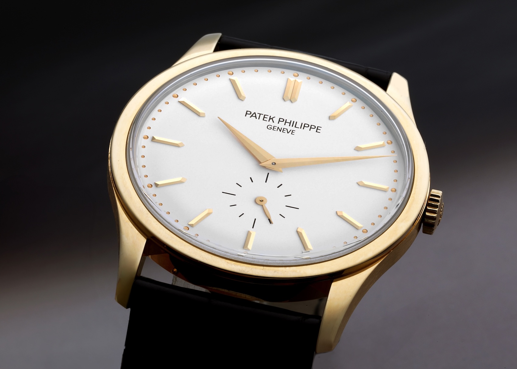 A close-up of a Patek Philippe Calatrava ref 5196J in yellow gold. The watch has a silver dial with gold baton hour markers, dauphine hands, and a small seconds sub-dial at 6 o'clock on a black strap.