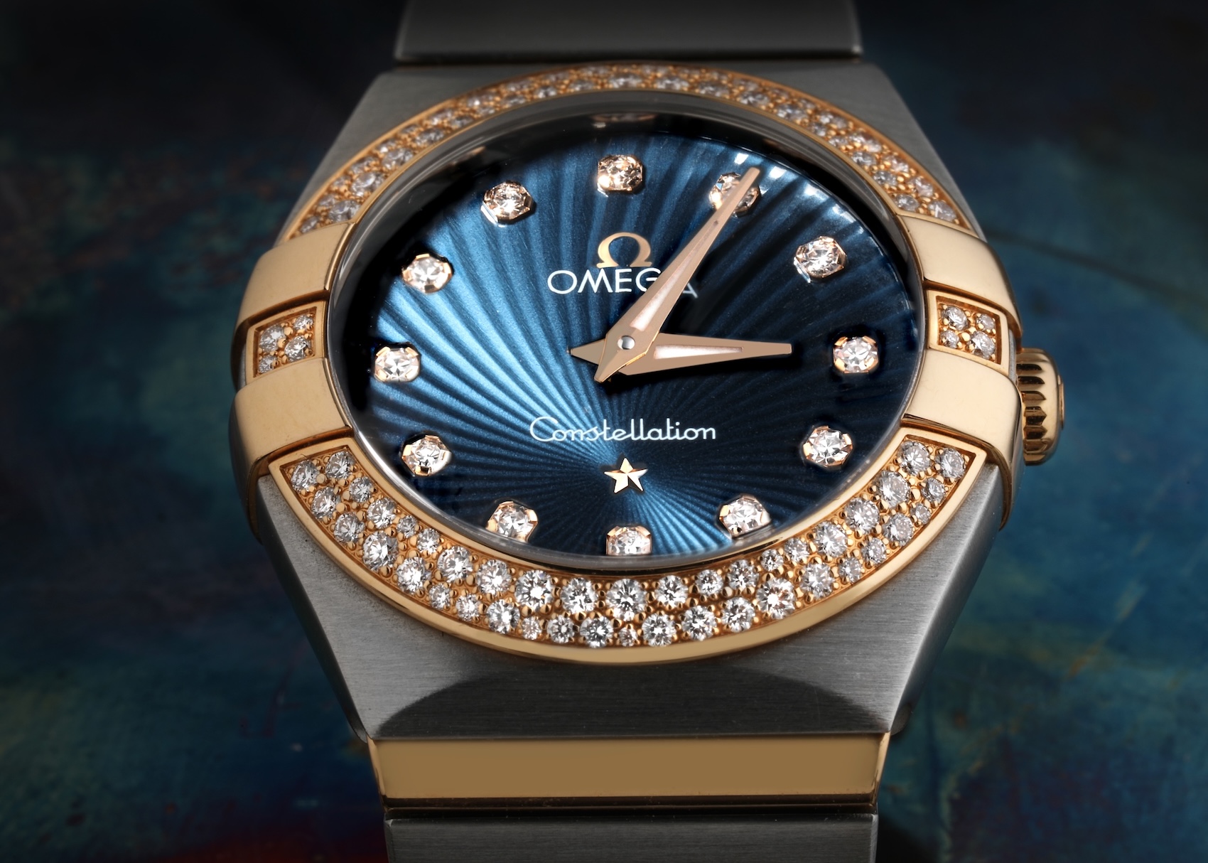 A detailed macro shot of an Omega Constellation ladies' watch, highlighting its diamond-paved bezel, textured blue dial with diamond hour markers, and two-tone steel and rose gold case.