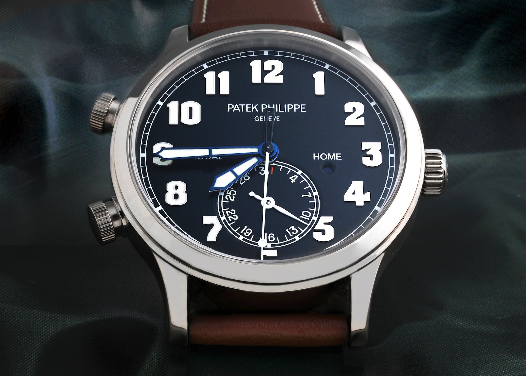 A head-on dial shot of the Patek Philippe Calatrava Pilot Travel Time 5524G, featuring a legible blue dial, large white gold numerals, and a brown leather strap.