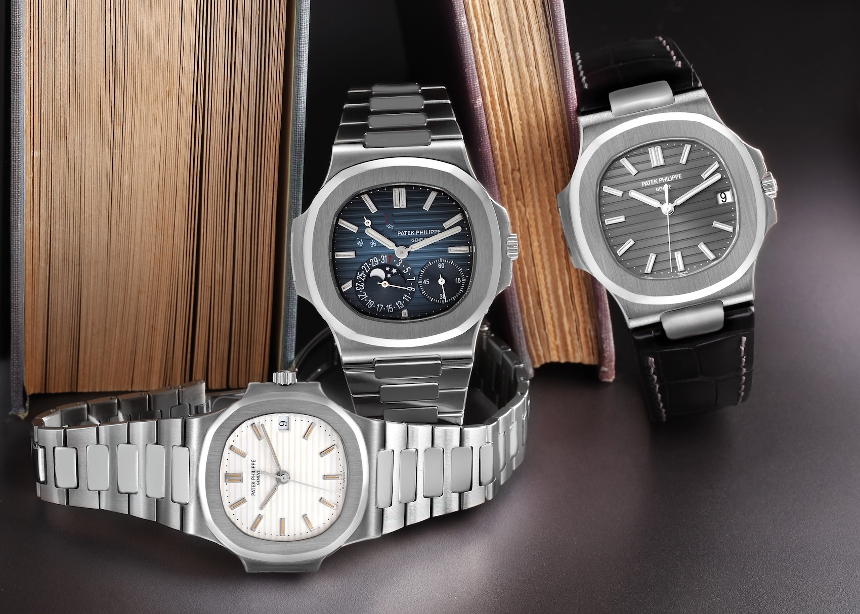 Three Patek Philippe Nautilus watches. In the foreground is a ref 3800 with a white dial. In the background are a ref 5712 moonphase with a blue dial and a ref 5711 on a leather strap with a grey dial.