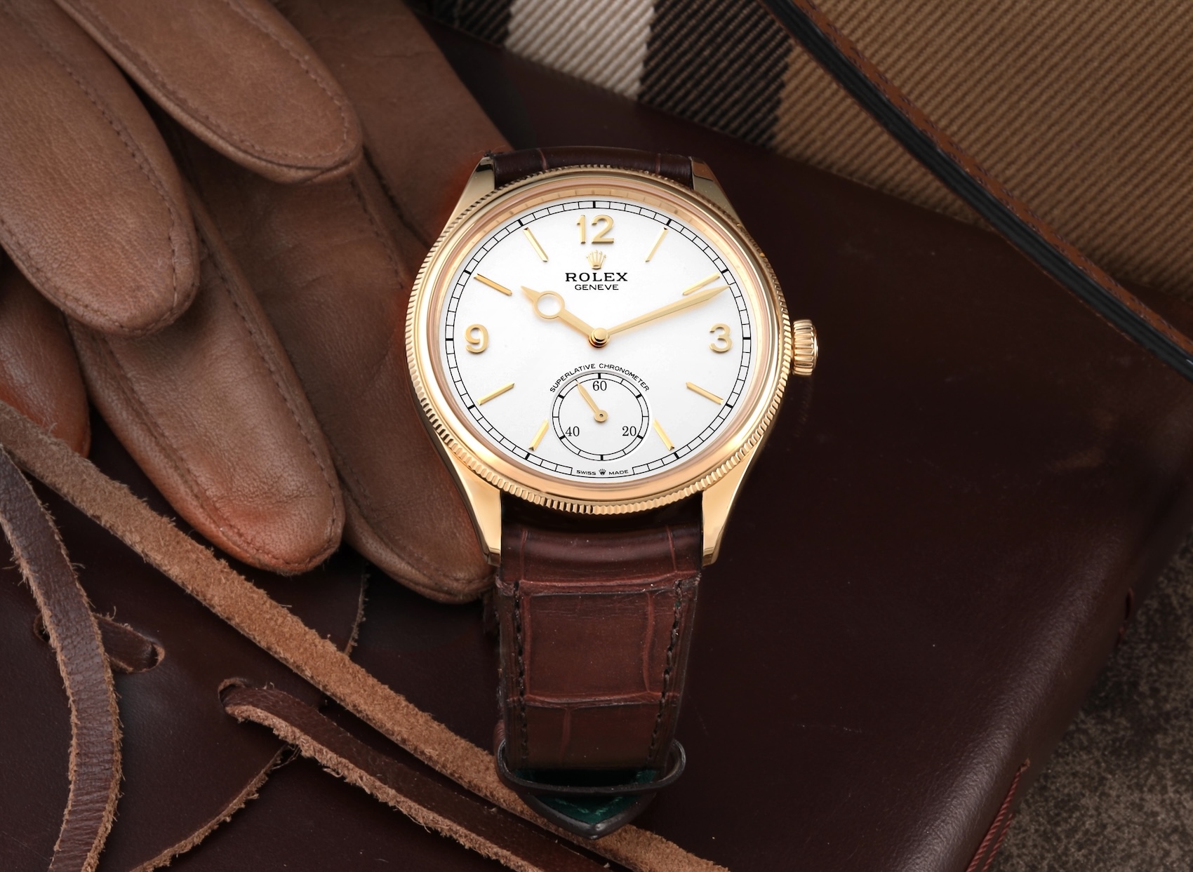 A close-up of the Rolex 1908 Perpetual dress watch, featuring an 18k yellow gold case, an intense white dial with a small-seconds subdial, and a brown alligator leather strap, styled with leather accessories.