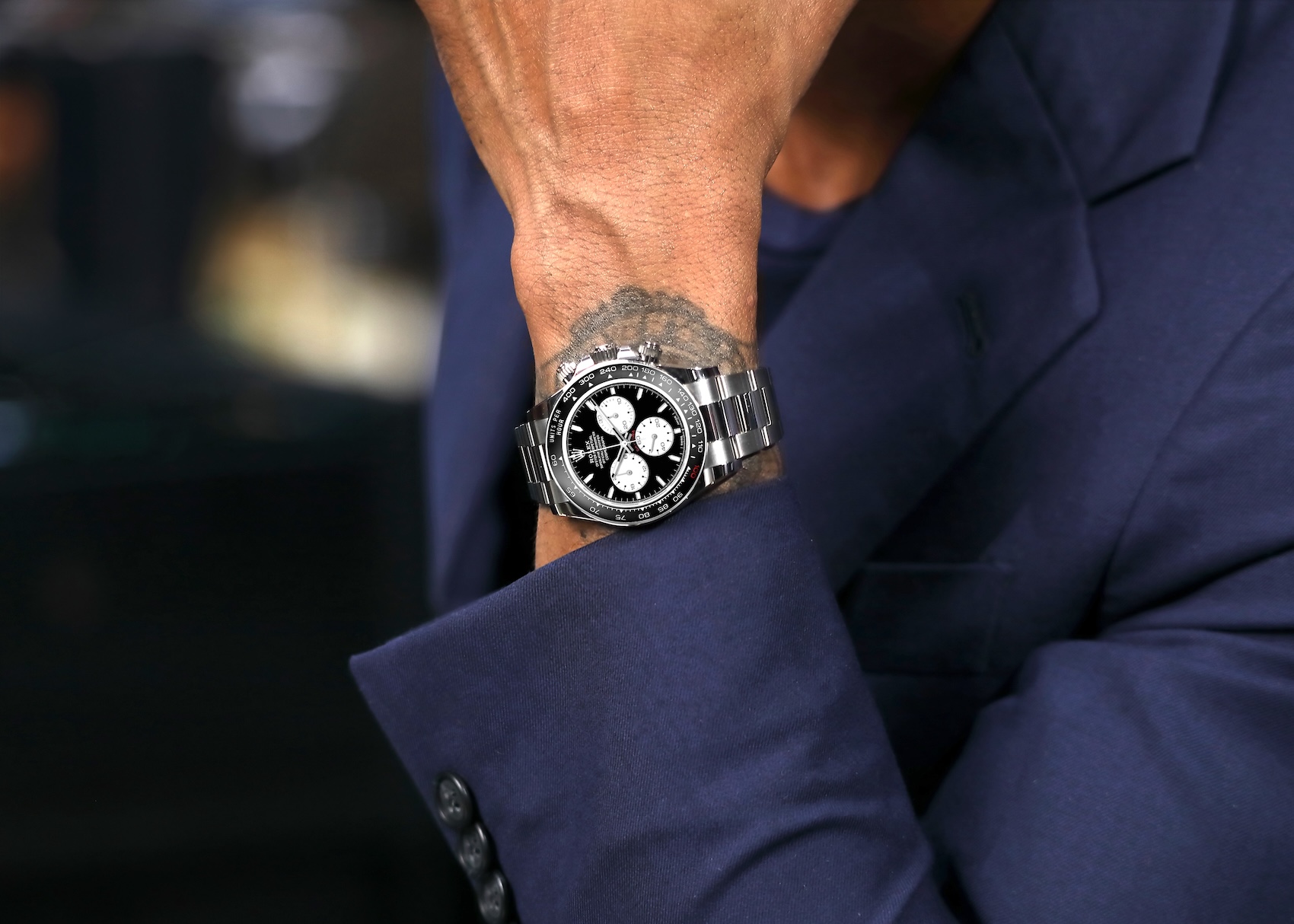 Rolex Daytona Le Mans watch worn on a wrist with a dark blue suit jacket sleeve visible.