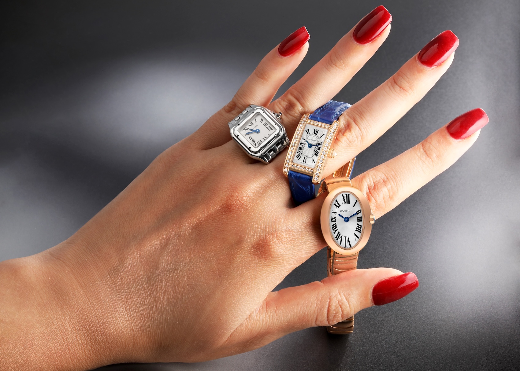 Hand modeling three mini Cartier watches: a steel Panthere, a diamond-set Tank Américaine on a blue strap, and a gold Baignoire.