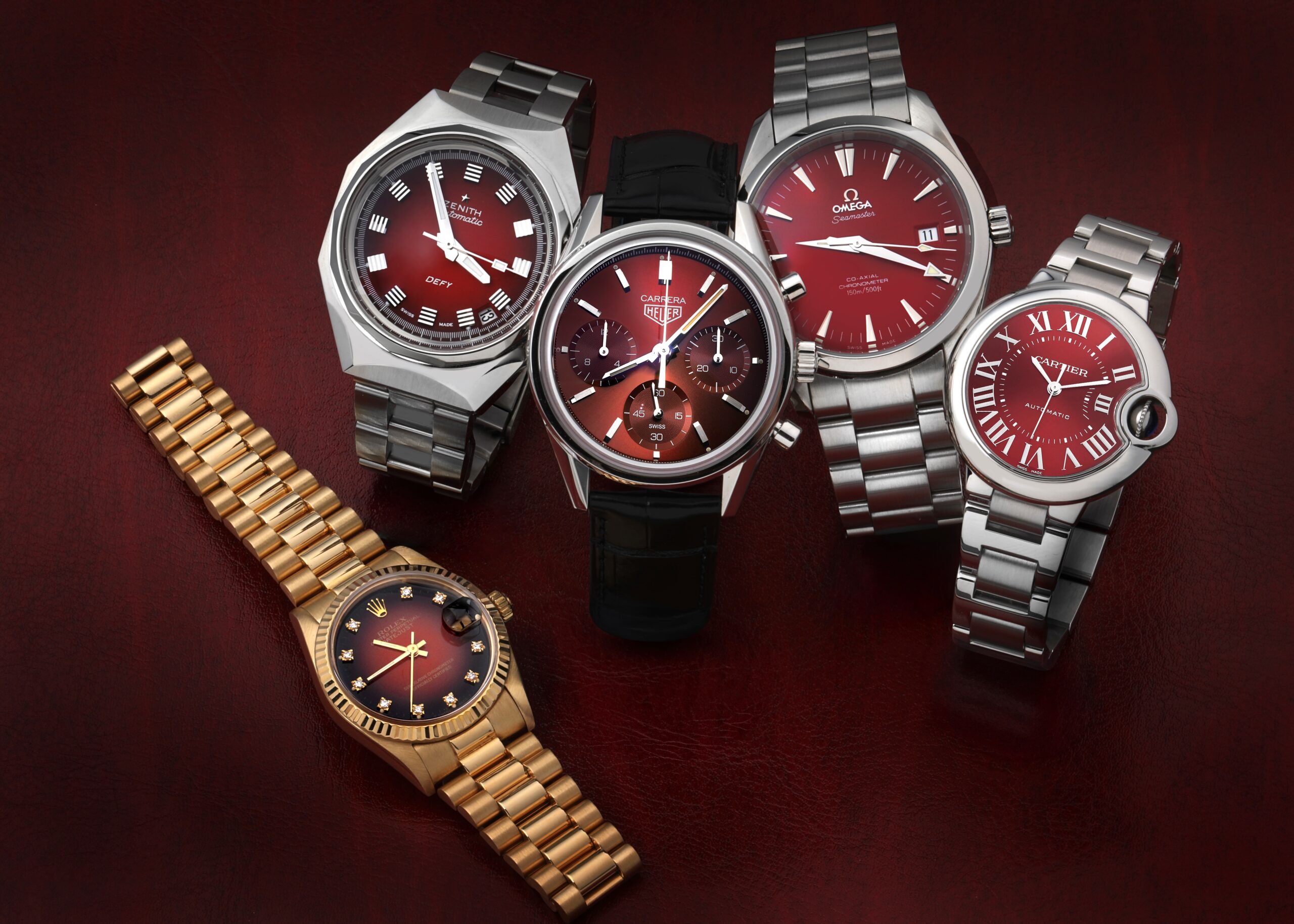 Collection of luxury watches with deep red dials including a gold Rolex Datejust, Zenith Defy Revival, TAG Heuer Carrera, Omega Aqua Terra, and Cartier Ballon Bleu, illustrating the Divine Damson color trend.