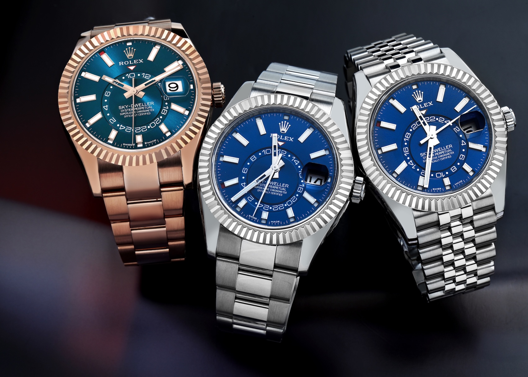 A group shot of three Rolex Sky-Dweller watches—one in Everose gold and two in Oystersteel—resting on a dark surface, showcasing the various bracelet and metal combinations.
