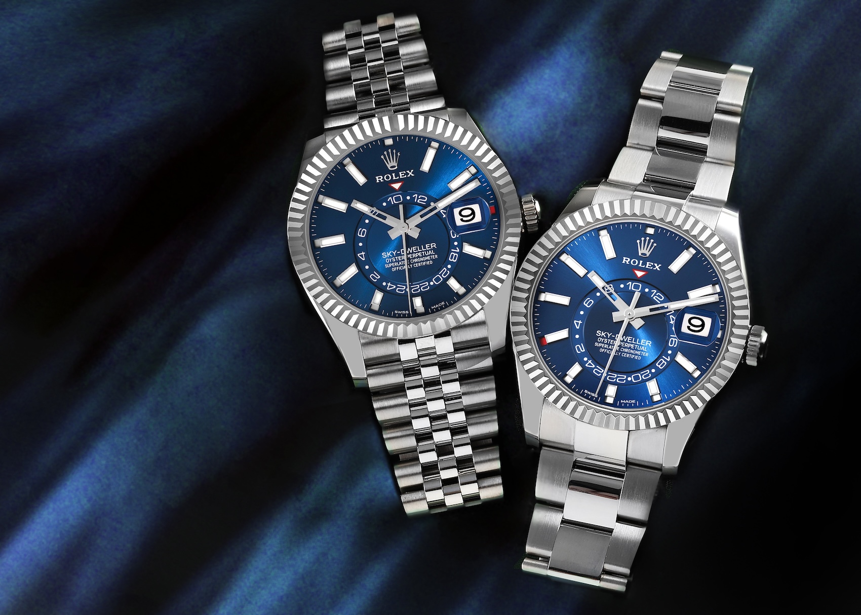Two Rolex Sky-Dweller 336934 watches with blue dials, one on a stainless steel Jubilee bracelet and the other on an Oyster bracelet, displayed on a dark blue fabric background.
