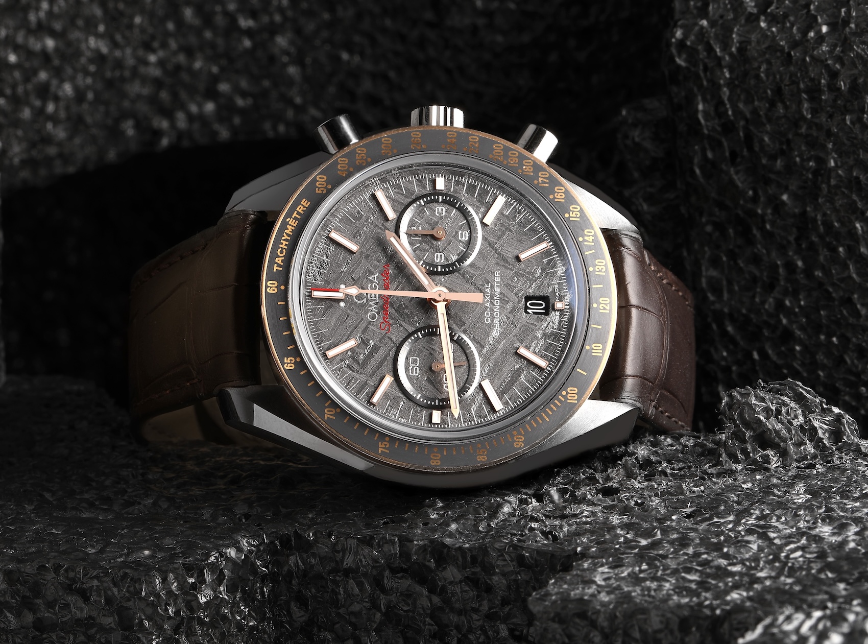 A luxury Omega Speedmaster "Grey Side of the Moon" watch featuring a full meteorite dial with a grey crystalline pattern, 18K Sedna gold hands and bezel, and a brown leather strap.