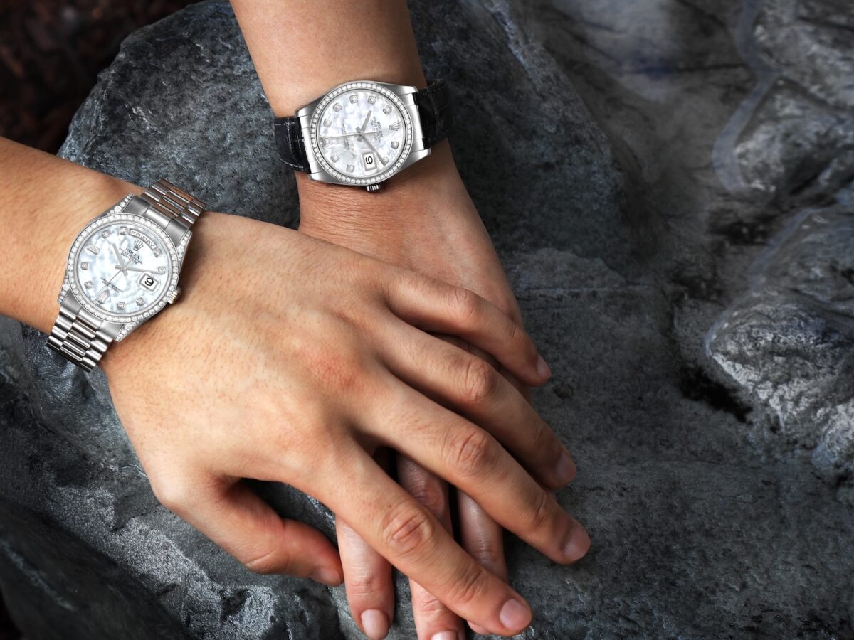 Top-down view of two wrists wearing a Rolex Day-Date 36 and a Rolex Datejust 36, both featuring Mother of Pearl dials and diamond bezels, resting on a dark grey rock.