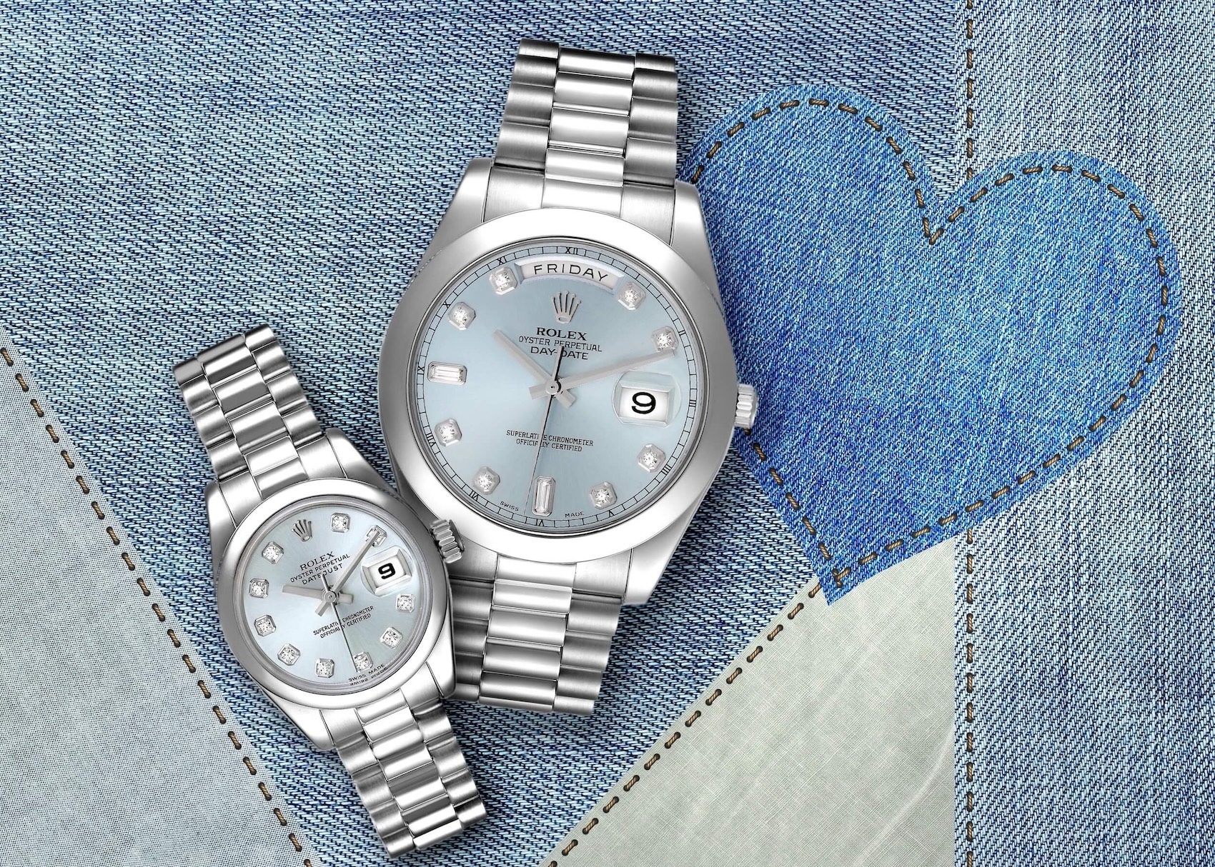 His and hers Rolex Platinum watches with ice blue diamond dials, arranged on a denim background with a heart design.