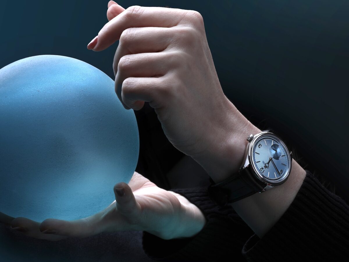 A moody, artistic photograph of a model holding a glowing blue frosted sphere, wearing the Rolex 1908 in platinum with an ice-blue dial on a brown leather strap.