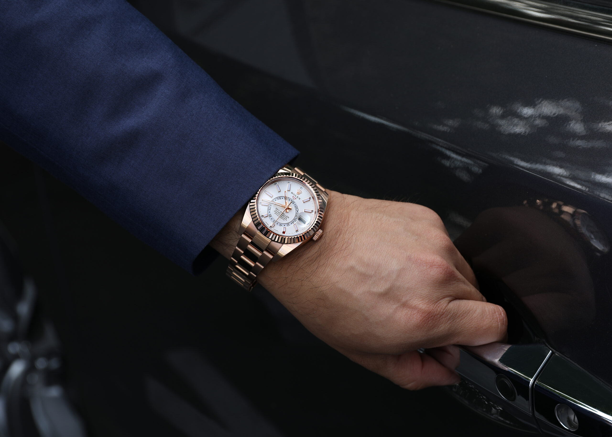 42mm Rose Gold Rolex Sky-Dweller on a man's wrist, resting on a luxury car door handle.