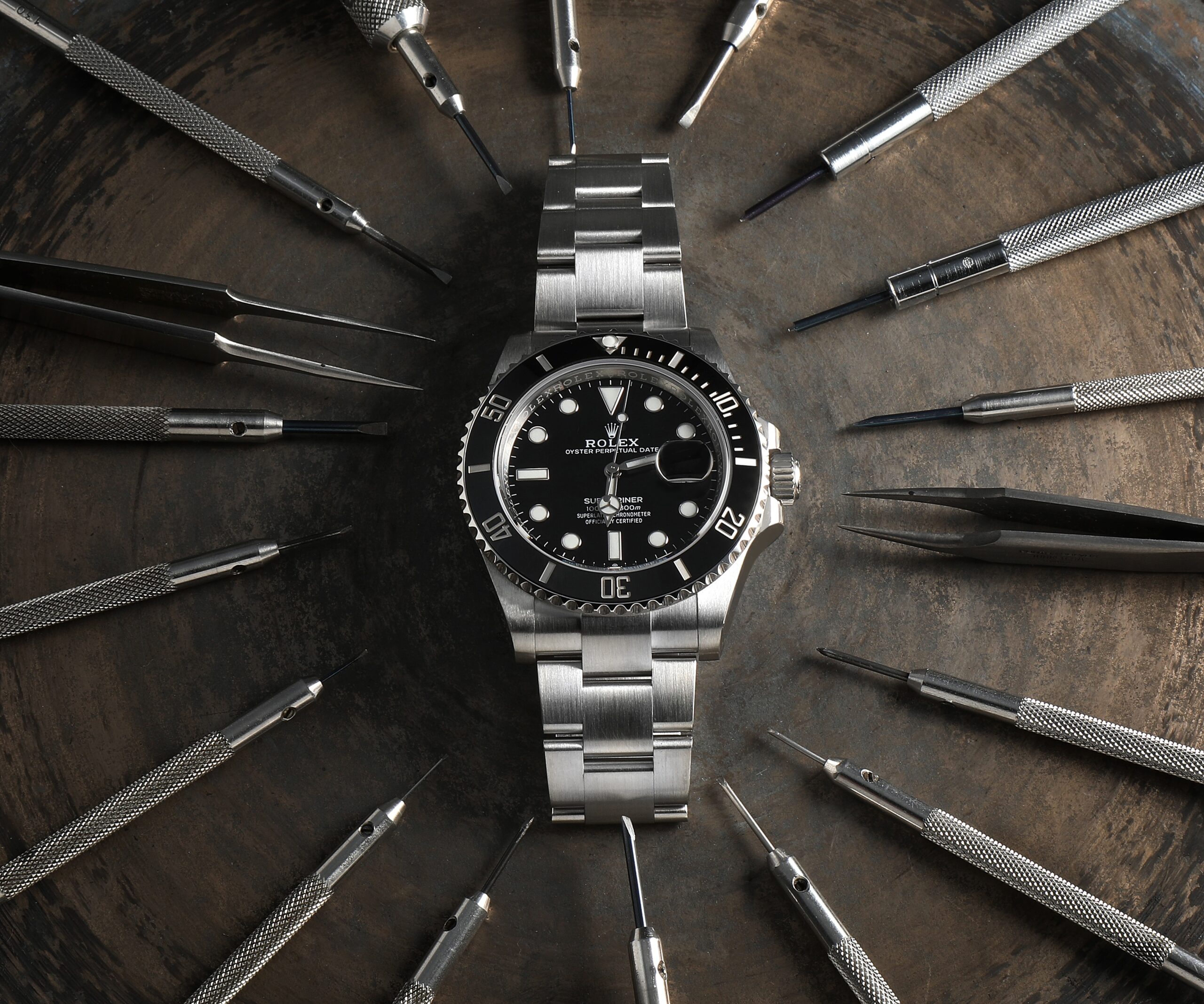 Top-down flat lay of a Rolex Submariner Date surrounded by a radial arrangement of silver watchmaker screwdrivers and tweezers on a dark surface.