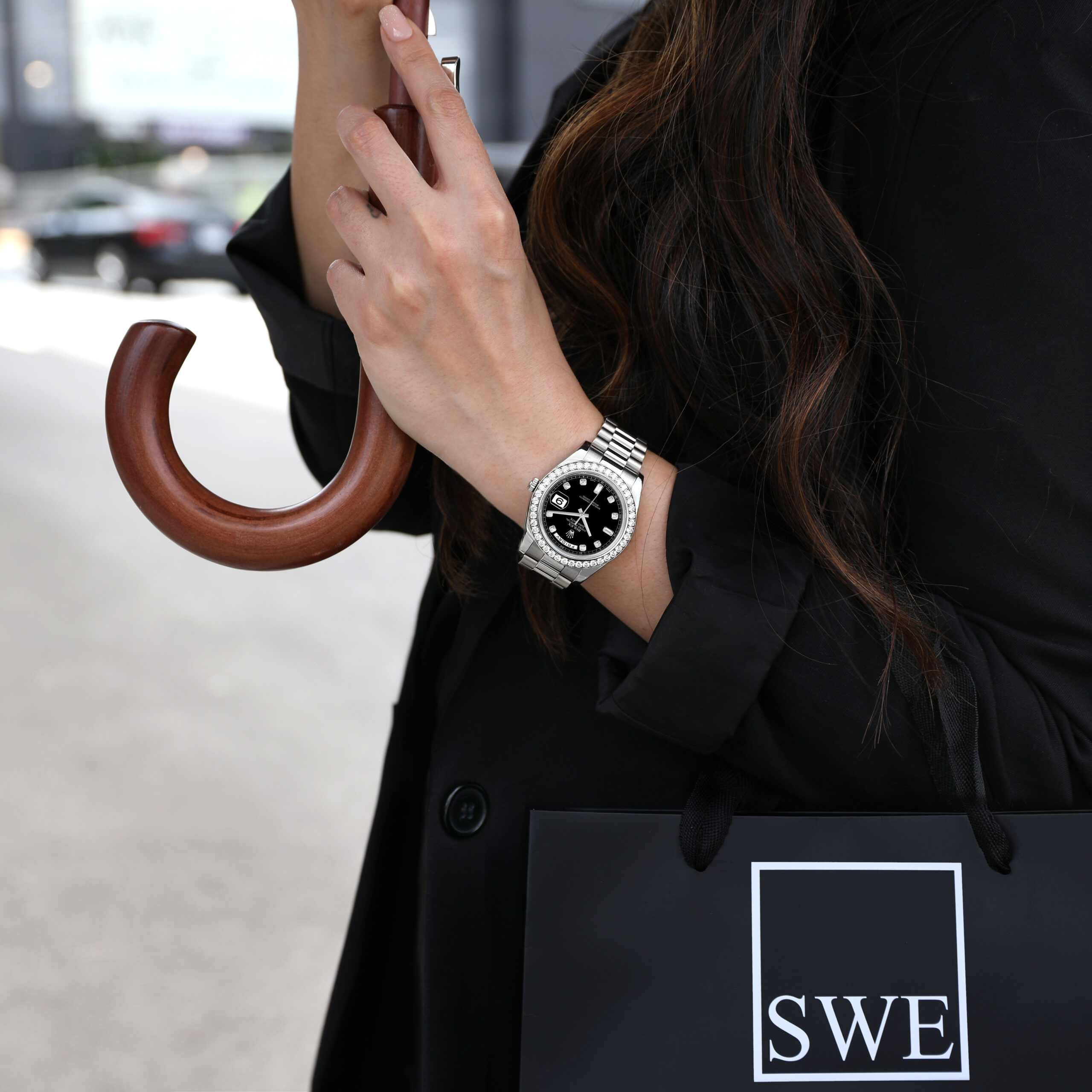 Woman wearing a stainless steel Rolex Day-Date with a diamond bezel, styled with a black blazer and umbrella.