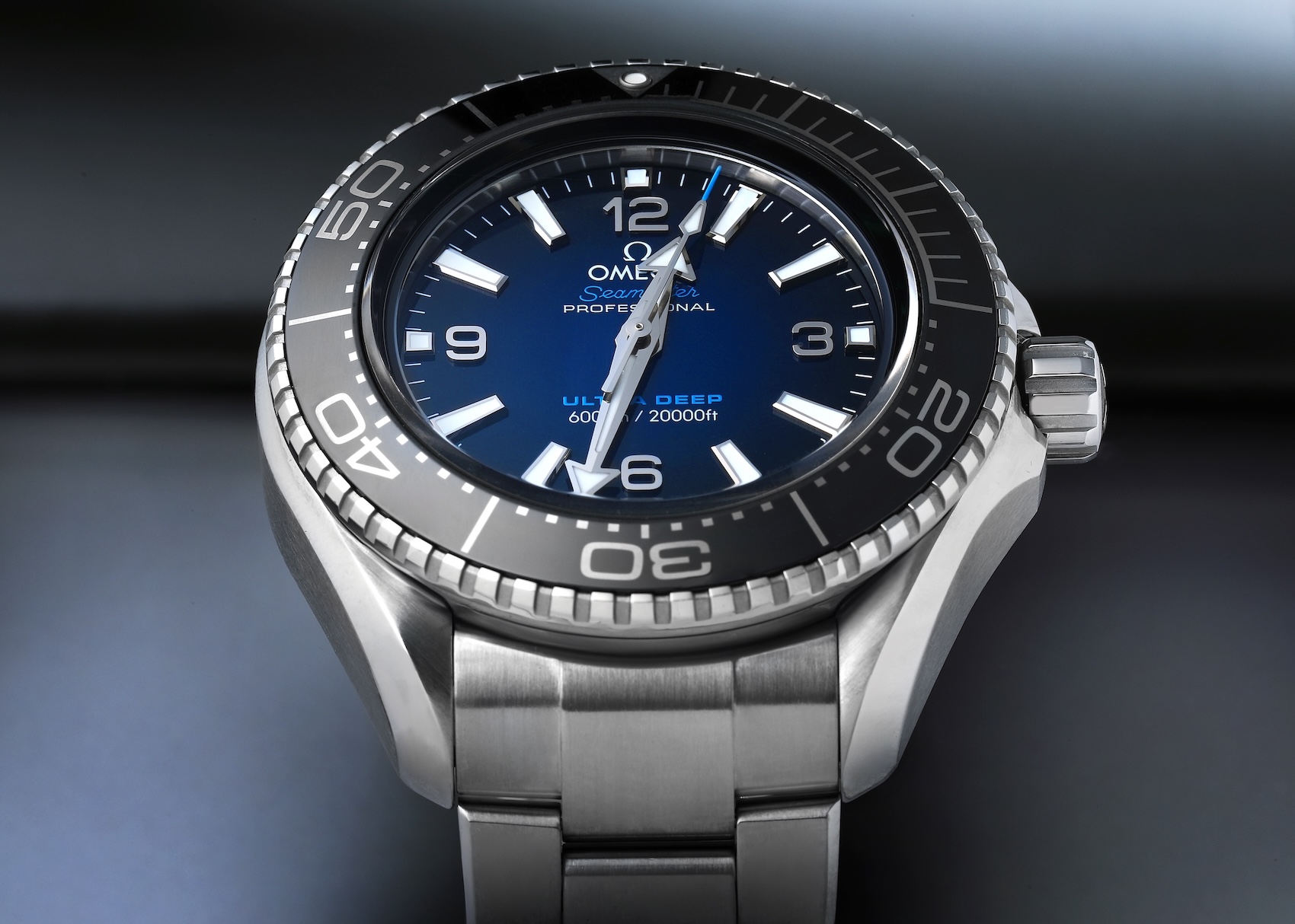 A sharp, angular perspective shot of the massive Omega Seamaster Planet Ocean Ultra Deep. It is made from high-performance O-MEGASTEEL and features a dark blue gradient dial, large indices, and a black ceramic bezel. It rests on its integrated steel bracelet.