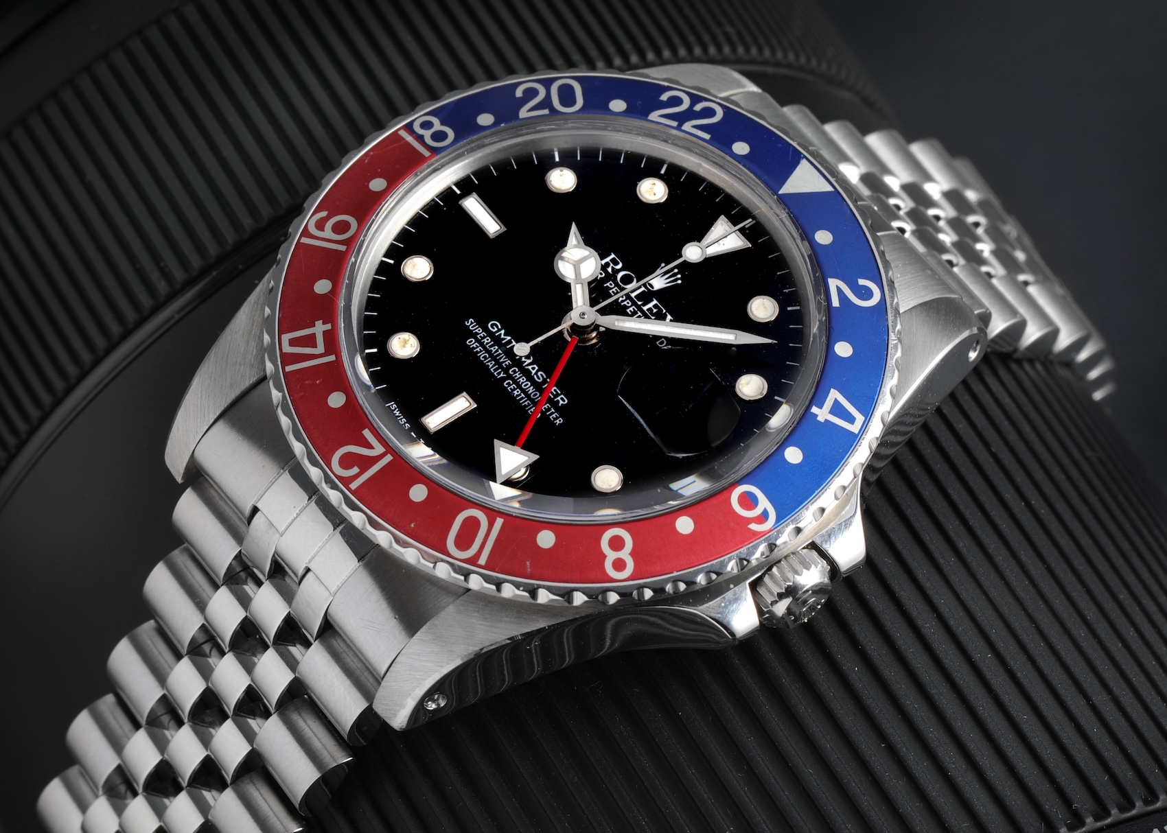 An angled close-up of a vintage stainless steel Rolex GMT-Master (Ref. 1675) with a faded red and blue aluminum Pepsi bezel, matte black dial, and a vintage Jubilee bracelet, resting on a black textured backdrop.
