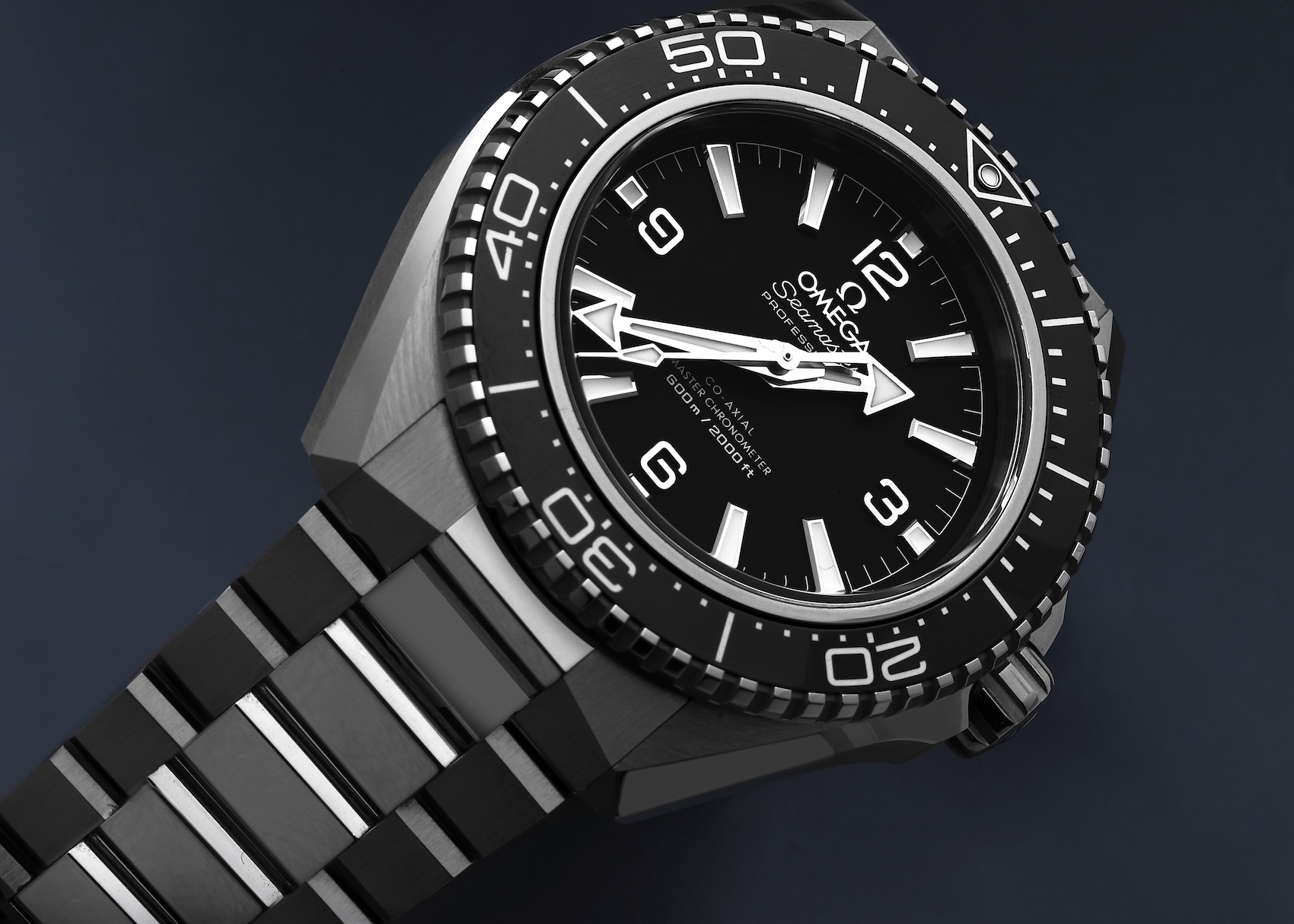 A dramatic, low-angle close-up photograph of the Omega Seamaster Planet Ocean 600M watch (ref. 217.30.42.21.01.001) set against a dark blue background. The shot highlights the sharp angular lines of the steel case, the deep black ceramic bezel, and the symmetrical 'no-date' dial with open-work Arabic numerals. The watch is shown on an unconventional dark, reflective bracelet with polished center links.