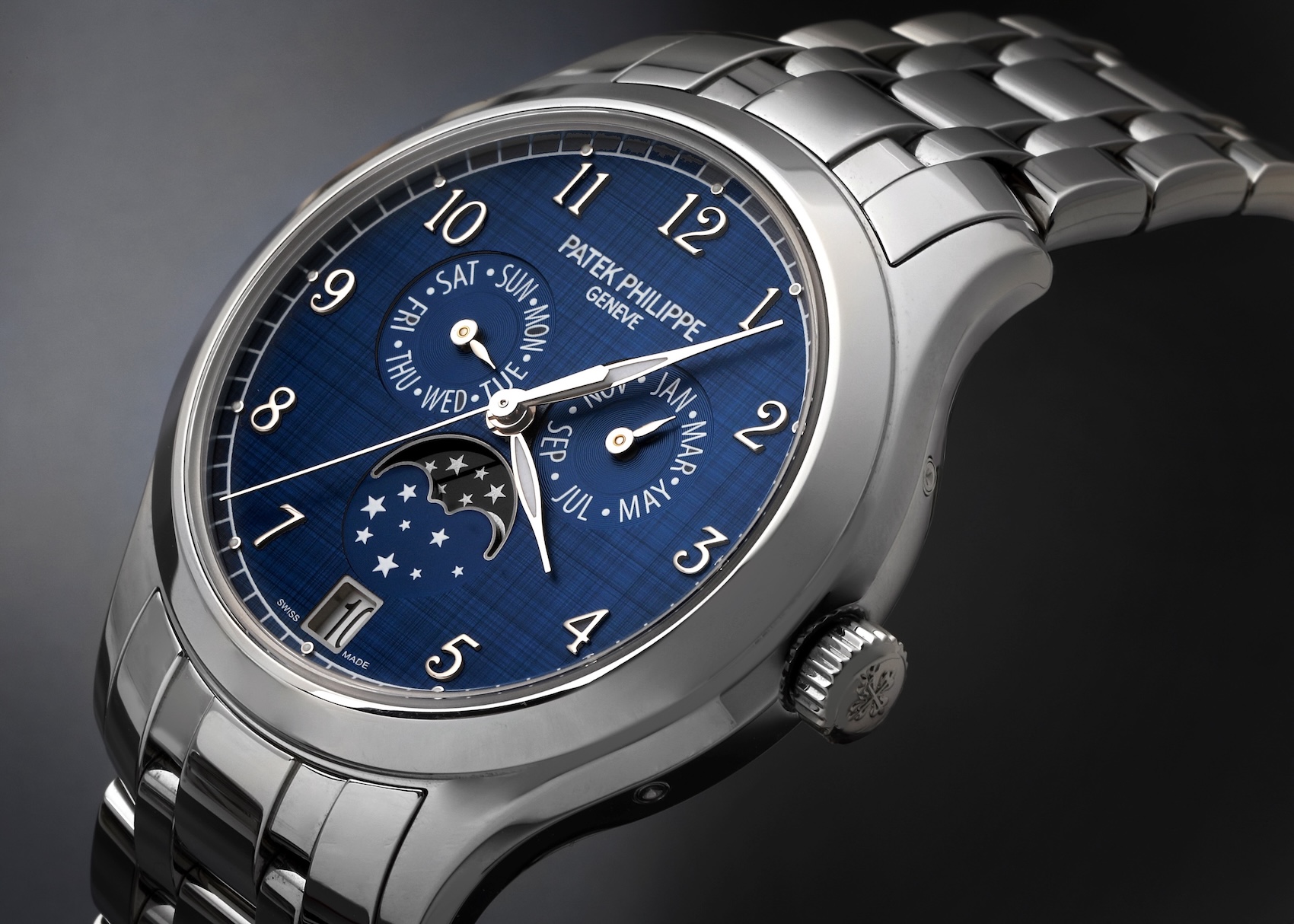 A close-up view of the dial of a Patek Philippe Annual Calendar watch. The dark blue dial features vertical and horizontal satin finishing, applied Arabic numerals, three subdials indicating day, month, and 24-hour time with a moon phase display, and a date window at 6 o'clock.