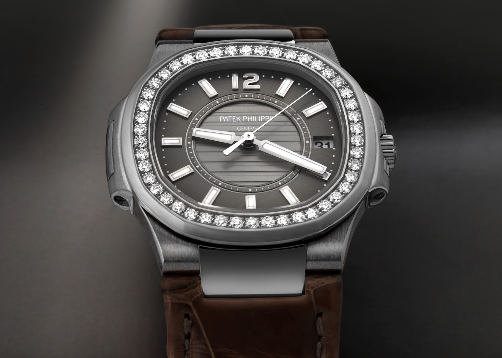A close-up view of the Patek Philippe Nautilus Ref. 7010G in white gold. The watch features a dark grey dial with the signature Nautilus horizontal embossing, a date window at 3 o'clock, luminous hands, and a bezel set with 46 brilliant-cut diamonds. It is paired with a brown alligator leather strap.